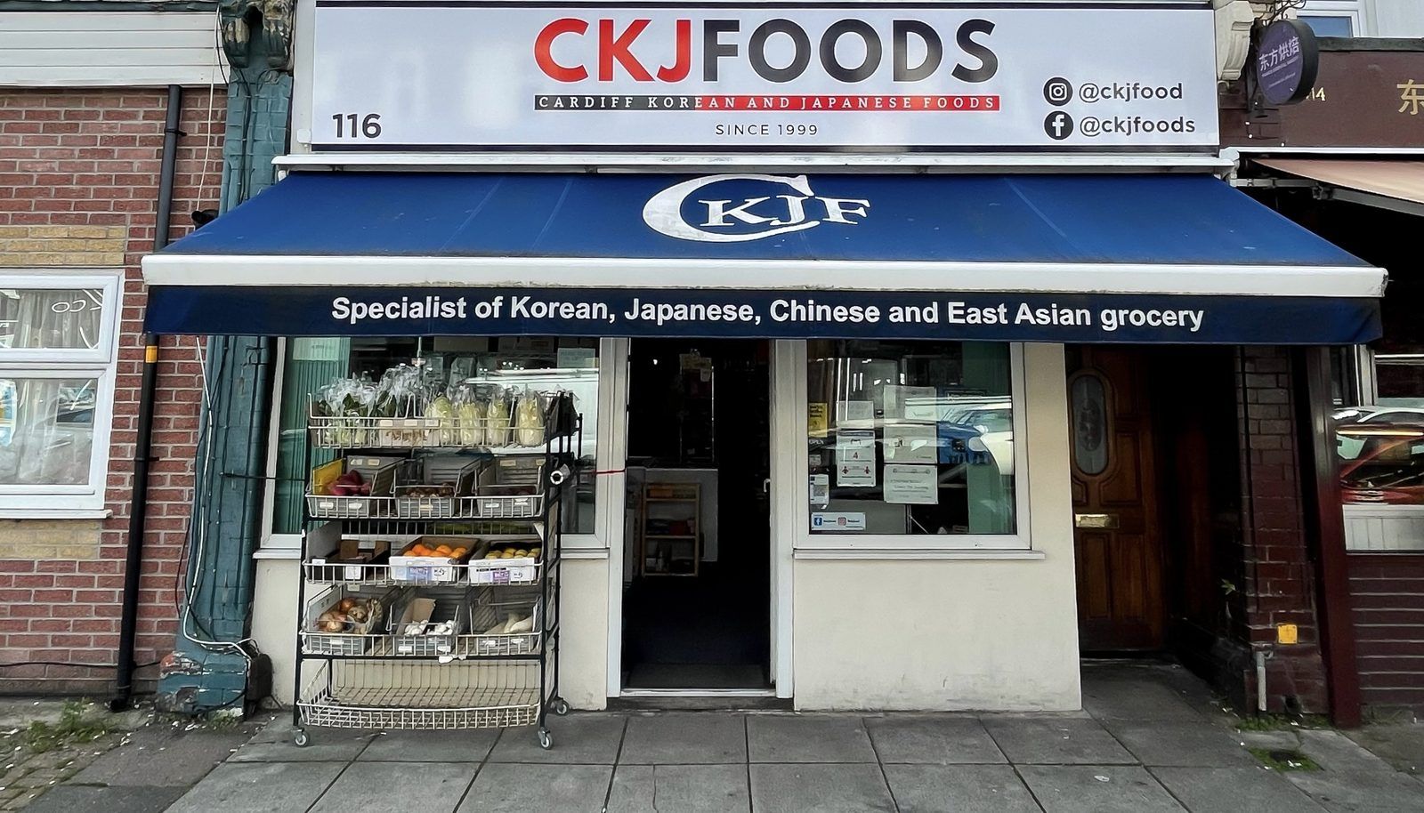 The CKJFoods shopfront at 116, with the red-and-navy CKJFOODS sign, blue CKJF awning, tagline banner and fresh produce displayed outside the entrance
