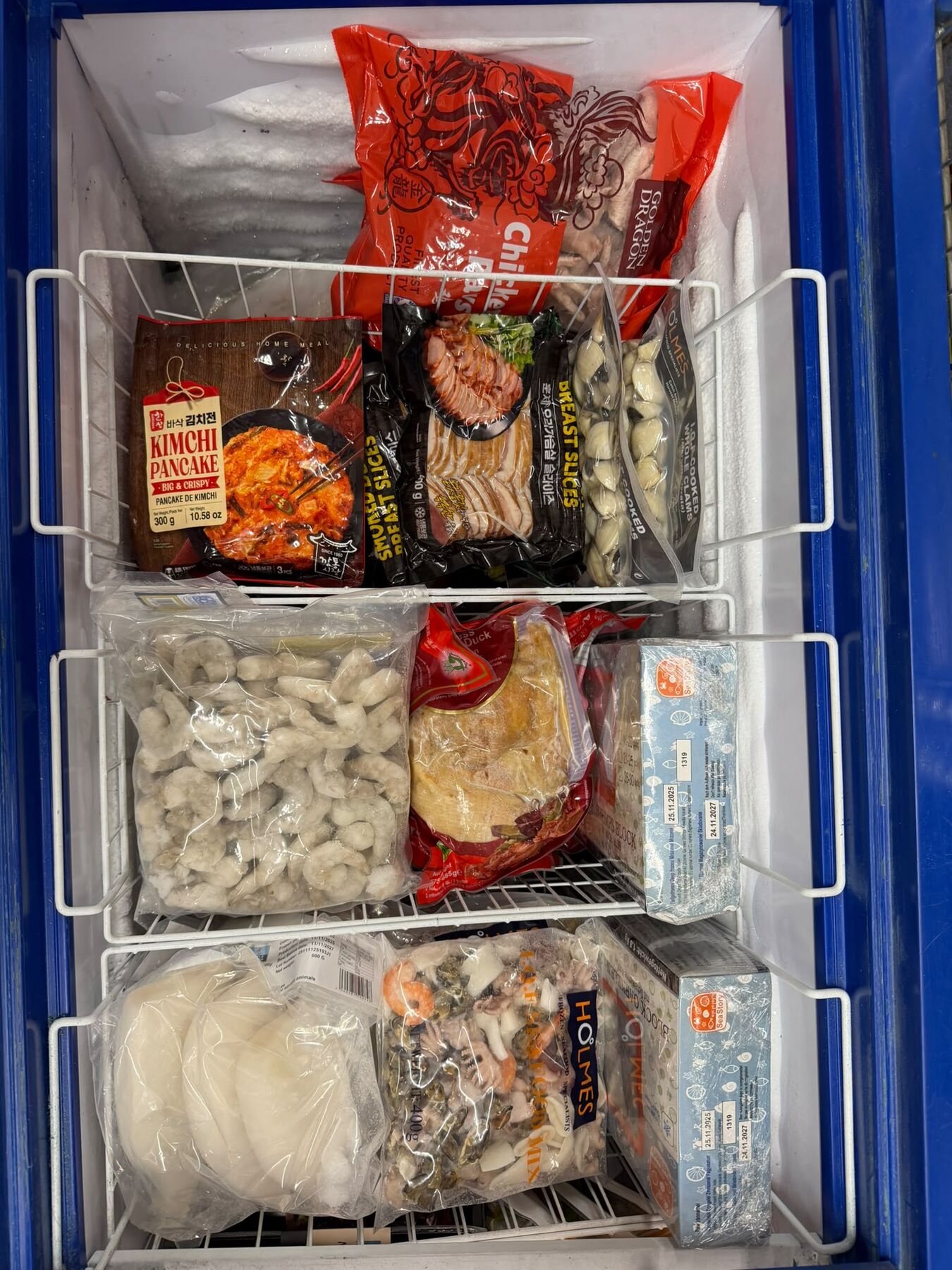 Freezer of frozen seafood and duck: kimchi pancake, smoked duck breast slices, cooked whole clams, chicken portions, raw peeled prawns, whole aromatic crispy duck, sliced rice cakes (tteok) and seafood mix